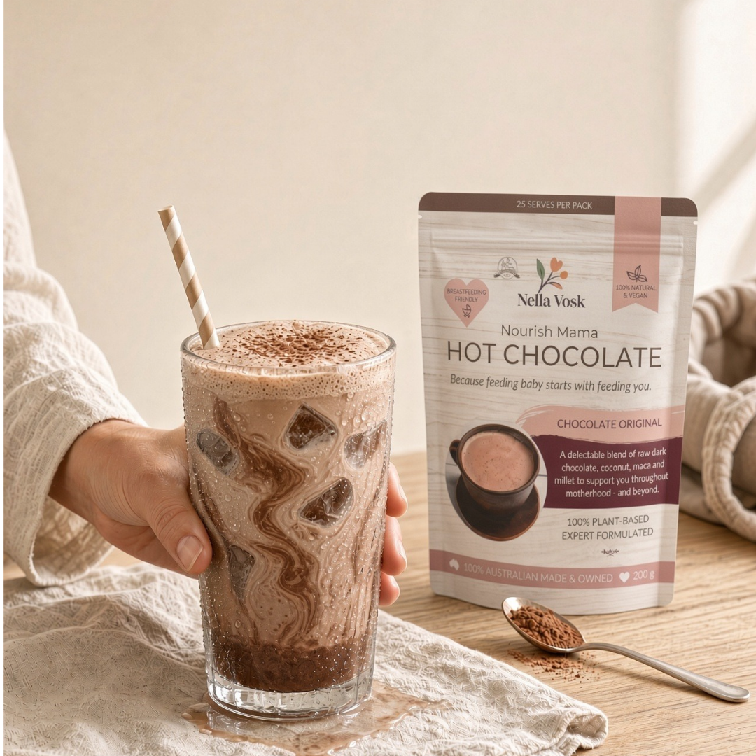 Dairy-free iced chocolate in a tall glass with ice, coconut whipped cream and a dusting of cacao, beside a pack of Nourish Mama Lactation Hot Chocolate, Nella Vosk